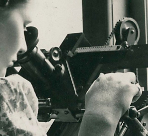 A black and white photograph of a woman looking through an eyepiece with a lens
